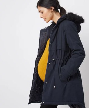 A pregnant woman wearing a navy faux fur trim hooded parka over a yellow jumper and black trousers.