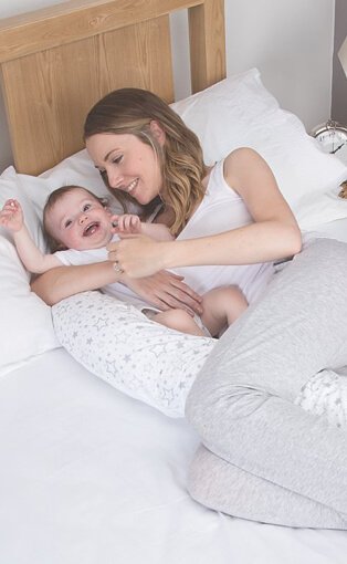 Mum wearing grey joggers and a white top lying in bed with a baby in a white bodysuit, using a maternity pillow for support