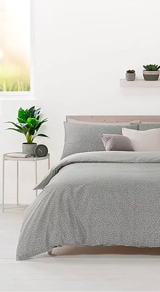 Double bed features green printed duvet set, mocha and cream scatter cushions with side table topped with artificial plant in white room with window in the background.