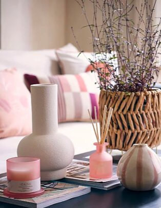 Cream living room features natural sofa with pink and cream scatter cushions with coffee table topped with artifical plant in wicker basket, cream vase, pink candle and reed diffuser set, selection of books and pink and cream stripey vase.