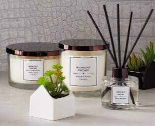 White surface topped with cream midnight orchid triple and double wick candles, midnight orchid reed diffuser and artificial plant in house-shaped vase.