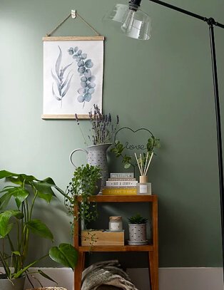 Green room features wooden table with assortment of books, artificial plants, heart-shaped ornament, candle and reed diffuser set, large jug with floor lamp to one side and leaf printed hanging frame in the background.