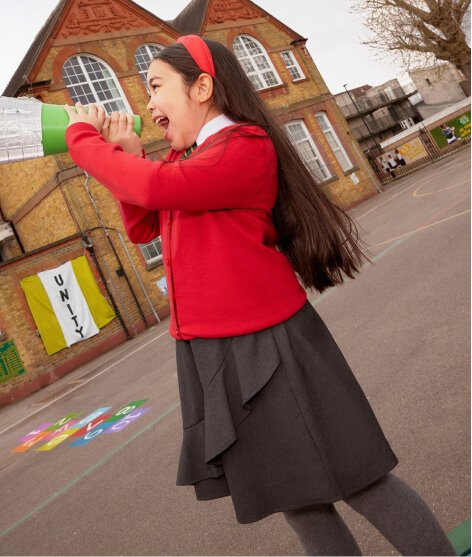 Girl in playground wearing red school jumper, grey pleated skirt and grey tights.
