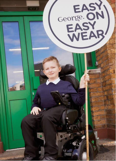 Boy smiles in a wheelchair wearing a white shirt, navy jumper and black trousers holding an 'easy on easy wear' sign.