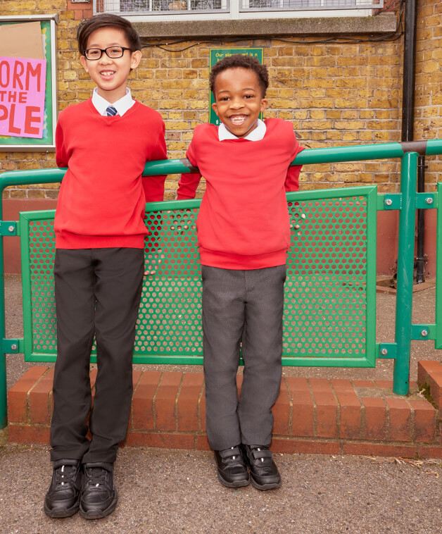 Two school children in front of a green railing wearing grey and black trousers, white shirts and red jumpers.