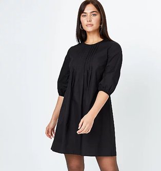 Woman poses wearing black pleated smock dress with ruched sleeves and gold-tone hoop earrings.