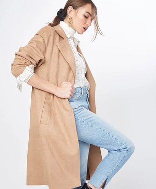 Woman poses side on raising one leg wearing cream high neck top, white checkered shirt, light wash blue jeans, tan faux suede belted throw-on jacket and gold-tone earrings.