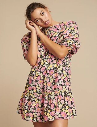 Woman poses resting head on hands wearing pink poplin floral balloon sleeve tiered mini dress and gold-tone hoop earrings.