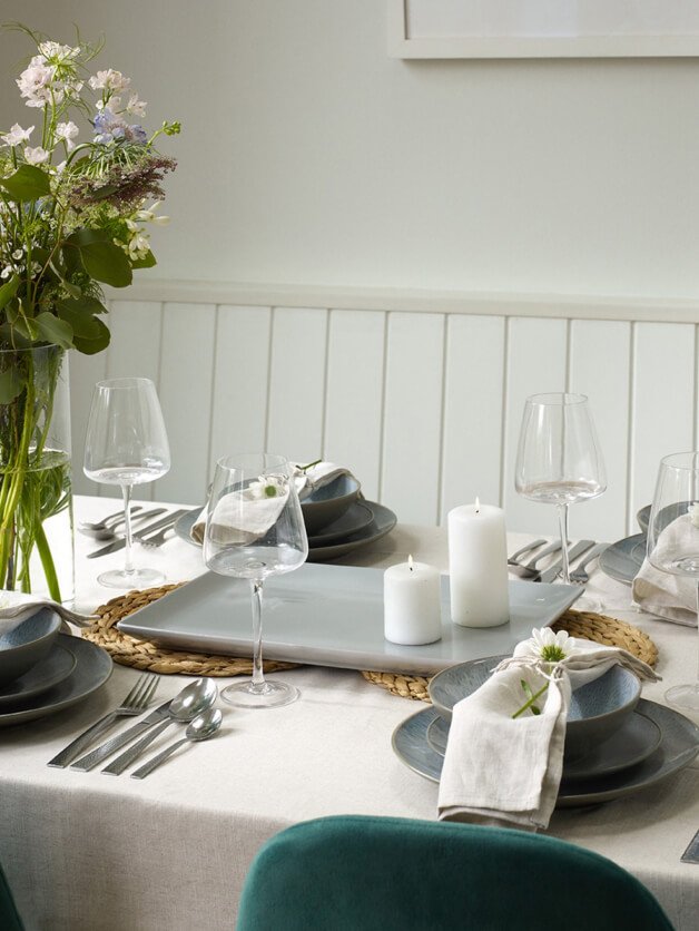A dining table topped with grey dinnerware, silver cutlery, wine glasses and decorative candles.