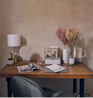 Pink room with wooden-effect table topped with open books, gold-effect white storage lamp, artificial plant, gold-effect storage box, white ribbed vase with pink artificial plant and cream and grey jug.