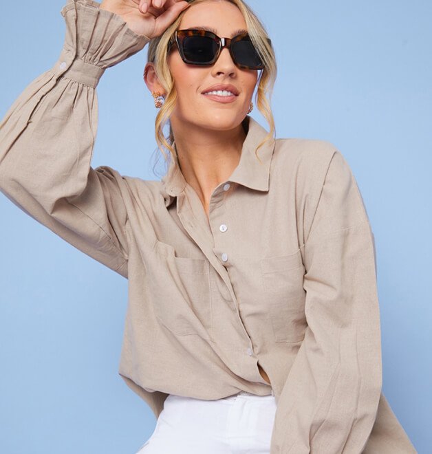 A woman posing in a beige blouse, white trousers and oversized sunglasses.