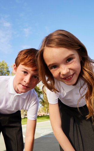 A boy wearing a white polo and black trousers standing in a playground with a girl wearing a white polo and a black skirt