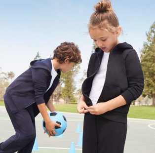 A girl and boy wearing navy joggers, zip up hoodies and white t-shirts, playing with a blue football in a playground