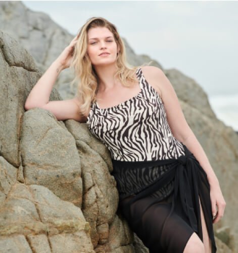 Woman poses on some rocks wearing a zebra print swimsuit and black sarong.