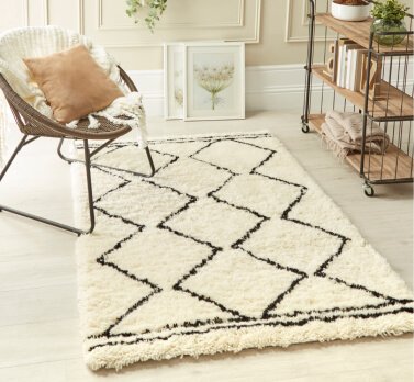 A living room with an armchair with a cushion and throw, monochrome geometric rug, wooden bookshelf and white framed prints.