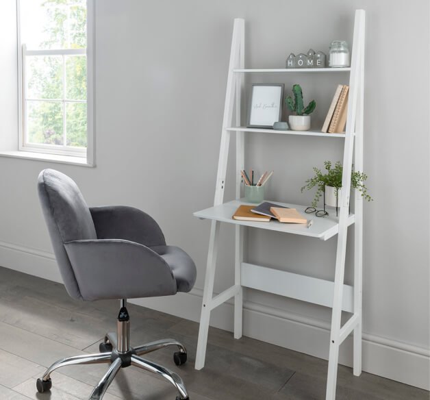 A grey office chair next to a white ladder desk topped with notebooks, pens, plants and other decor items.