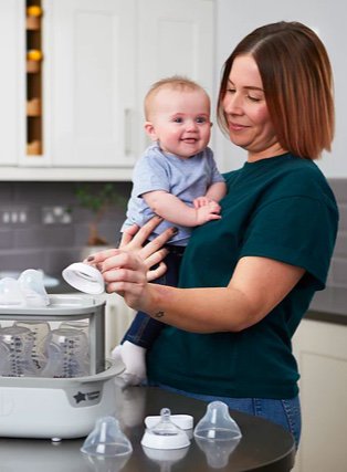 Woman stands in kitchen sterilising baby bottles holding baby boy in her arms.