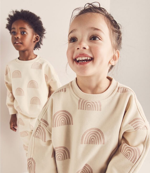 Two kids smiling in matching cream rainbow jumper and jogger outfits.