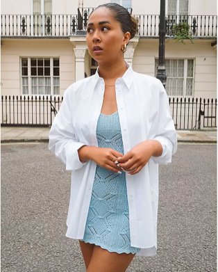 Woman poses looking away from camera wearing white oversized shirt over blue mini dress.