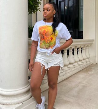 Woman poses with hand in pocket wearing white sunflower graphic t-shirt tucked into white fray hem shorts with white trainers.