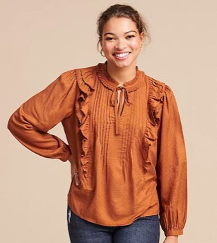 Woman poses smiling with hand on hip wearing tan textured frill tie neck blouse.