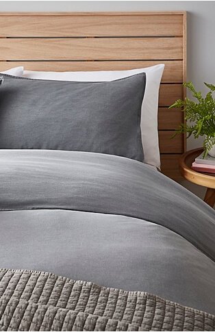 Section of bed with wooden headboard, grey bedding and textured grey throw with wooden bedside table with plant. 