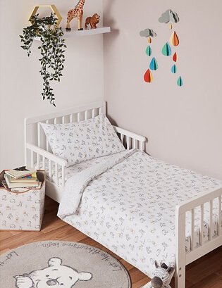 A toddler bed with white Winnie The Pooh bedsheets in a bedroom with a matching storage box and circular rug, white shelf with toys and a raindrop wall decoration.