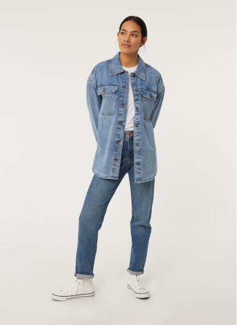 A woman posing in a denim jacket, jeans, white t-shirt and trainers.