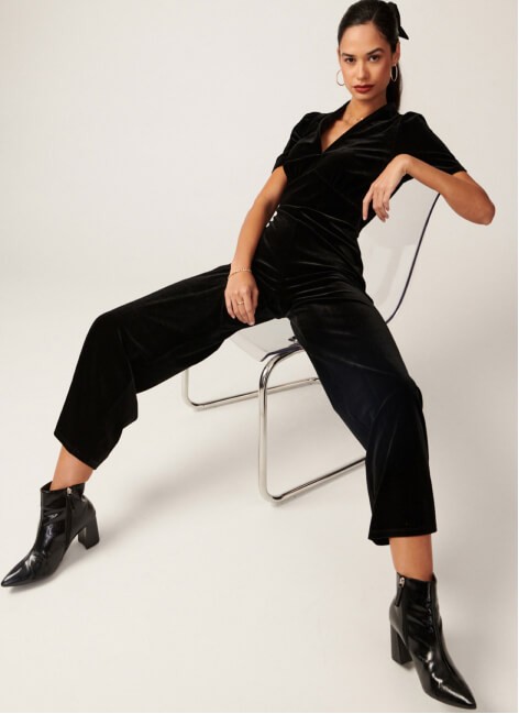 A woman posing in a black velvet v-neck jumpsuit and black boots.