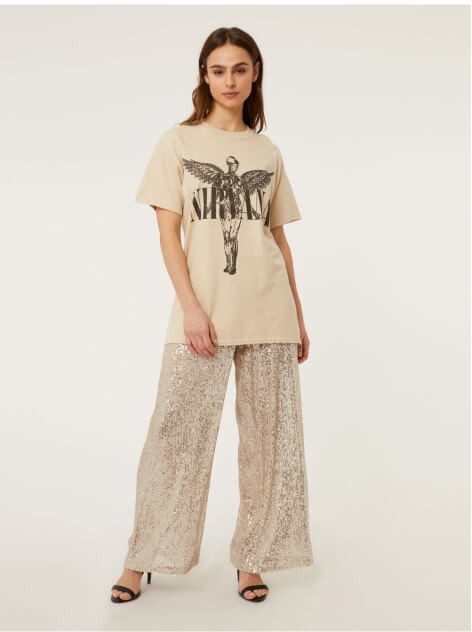 A woman wearing a Nirvana cream band t-shirt, cream sequin trousers and black heeled sandals.