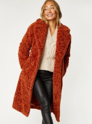 Woman wearing rust coloured fluffy coat, cream top and black faux leather trousers.