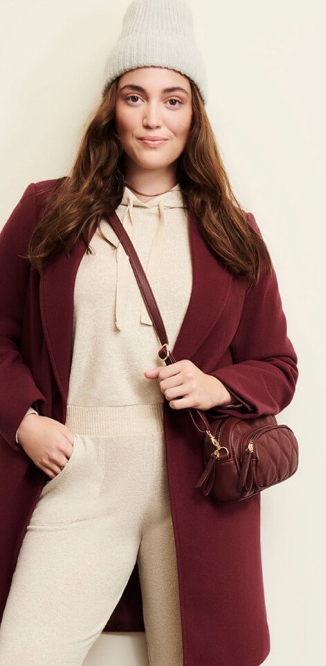 A woman wearing a neutral soft touch set, a plum longline coat, grey beanie and a cross body bag.