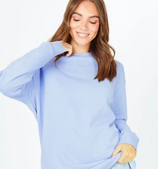 Woman poses raising hand to neck and holding hem of blue sweatshirt.