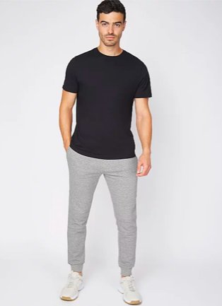 Man poses wearing black t-shirt, grey marl jersey joggers and white trainers.