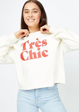 Woman poses smiling wearing cream très chic slogan sweatshirt and blue light wash jeans.
