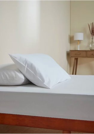 Cream room features double bed with white sheet and pillows with wooden dresser in the background topped with cream lamp.