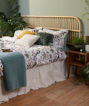 Double bed with floral bedding, green, cream and yellow scatter cushions, cream valance, green textured throw with wooden side table. 