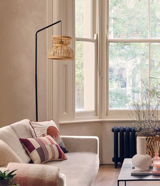 White room with open windows features cream sofa with multicoloured scatter cushions, black floor lamp with wicker shade and coffee table.