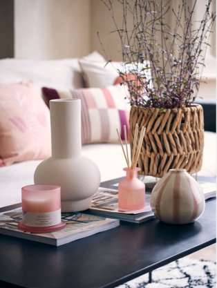 Cream sofa with pink, beige and cream scatter cushions, matte black coffee table with large wicker artificial plant, white vase, pink and white stripe vase, pink candle and reed diffuser.