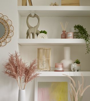 White shelf with giraffe ornament, rainbow artificial plant, pink candle and diffuser and artificial trailing plant, white shelf with bamboo lamp, pink candle, white vase and elephant artificial plant next to wall with round mirror and pink artificial plant in white vase.