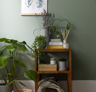 Wooden table topped with grey jug vase, 3 stacked books, reed diffuser, love wire sign and artificial trailing plant next to large artificial plant in grey pot.