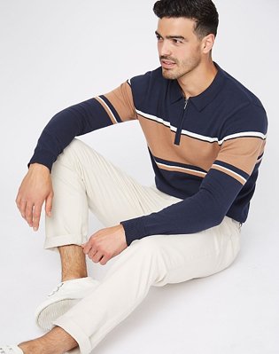 A man sitting on the floor wearing a navy striped zip polo jumper with cream trousers and white trainers.