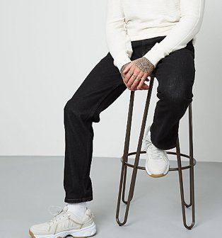 A man sitting on a bar stool wearing black straight fit jeans, a white jumper and white trainers.