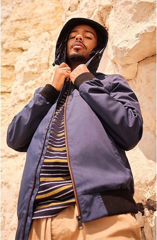 Man posing in front of a large rock wall wearing a navy bomber jacket over a navy and yellow stripe t-shirt and tan trousers.