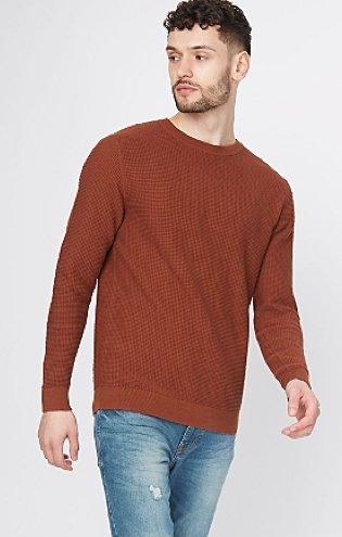 A man looking to the side wearing a brown lightweight textured crew neck jumper and distressed blue jeans.