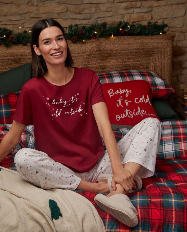 A woman sitting on a festive bed wearing a 'Baby it's cold outside' slogan pyjama set