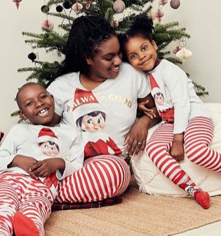 Woman, a young boy and a young girl sit cuddled in front of a Christmas tree wearing matching family Christmas Elf on the Shelf pyjamas.