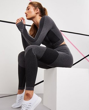 A woman sitting on a white box wearing grey WRKOUT leggings and long sleeve top with white trainers.