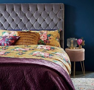 A double bed with a yellow floral bedspread, fringe accent cushions and a plum throw next to a bedside table with a glass lamp and a vase of pink flowers.