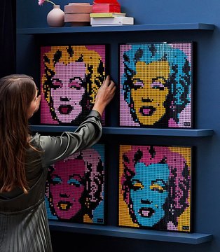 A woman placing four LEGO Art Andy Warhol portraits on a wall shelf.